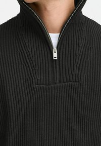 Gros plan d'un pull en maille côtelée noire avec une fermeture éclair argentée partiellement ouverte, porté sur un sous-vêtement blanc.