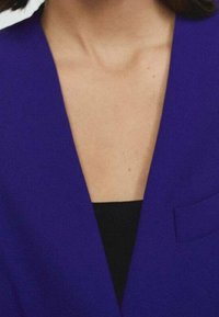 Fioletowa marynarka z głębokim dekoltem w serek, gładką teksturą materiału i małą kieszonką na piersi, noszona na czarnej bluzce.