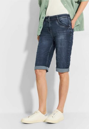 Person iført opkørte mørke denim knælange shorts, hvide sneakers, hvid top og grøn skjorte med knapper med hænderne i lommerne.