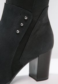 Bottine en suède noir avec un haut talon bloc, dotée de trois boutons décoratifs en argent sur le côté extérieur et de détails de couture.