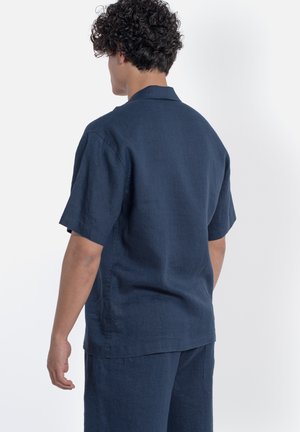 Uomo con capelli ricci che indossa una camicia blu navy a maniche corte e pantaloni abbinati, visto da dietro su sfondo bianco.