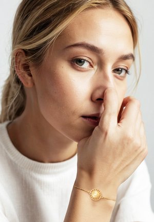 Guldarmbånd med en rund vedhæng, der har et radialt mønster, båret på en hånd nær en models ansigt, sat mod en neutral baggrund.