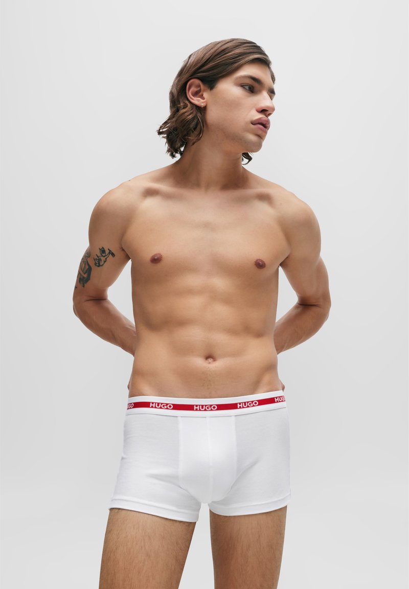 Boxers en coton blanc au design ajusté avec une ceinture rouge "HUGO". Le modèle se tient avec les bras derrière, mettant en valeur un torse musclé.