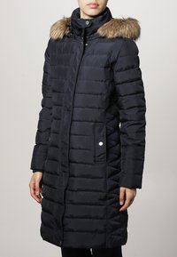 Marine blauwe gewatteerde jas met een afneembare faux fur capuchon, gestepte design, zakken aan de voorkant en een ritsluiting. Gladde textuur met een strakke pasvorm.