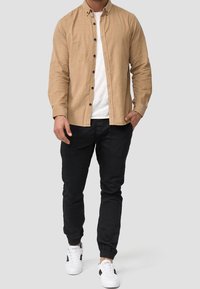 Hellbraunes Hemd zum Knöpfen über einem weißen T-Shirt, kombiniert mit schwarzen Jogginghosen und weißen Sneakern mit schwarzen Akzenten.