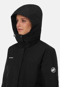 Giacca nera impermeabile con cappuccio, dotata di chiusura con zip e bottoni a pressione. Logo Mammut ben visibile sul petto e sulla manica. Texture liscia.