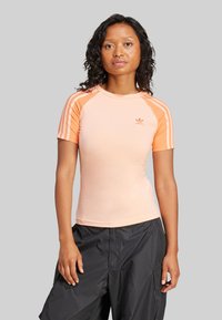 Persikka kappa lyhyillä hihoilla, jossa on valkoiset vaakasuorat raidat hartioilla ja pieni logo rinnassa. Sileä tekstuuri, slim fit -malli.