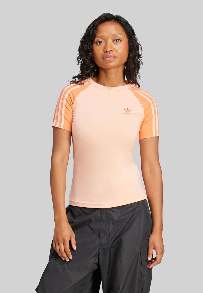 Persikka kappa lyhyillä hihoilla, jossa on valkoiset vaakasuorat raidat hartioilla ja pieni logo rinnassa. Sileä tekstuuri, slim fit -malli.