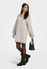 Jasnoszary, oversize, dzianinowy sweter typu sukienka z długimi rękawami, ściągaczowymi mankietami i okrągłym dekoltem, w zestawieniu z czarnymi botkami za kostkę i małą torebką.