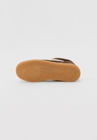 Baskets en suède marron avec une semelle en caoutchouc gomme. La semelle présente un motif alvéolaire pour une meilleure traction. La tige en tissu doux comprend un accent de couleur crème.
