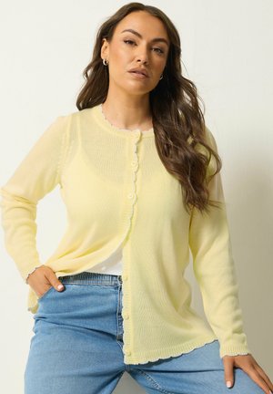 Kvinde med langt brunt hår iført en lys gul knappecardigan og blå jeans, poserende med den ene hånd i lommen mod en neutral baggrund.