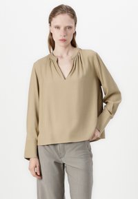 Blouse beige à manches longues avec décolleté en V, détail de col froncé et coupe décontractée. Associée à un pantalon droit gris clair.