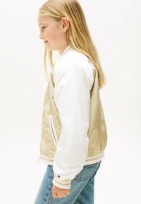 Chaqueta bomber beige y blanca hecha de tejido brillante, con cremallera, bolsillos laterales y puños a rayas. Combinada con jeans de color azul claro.