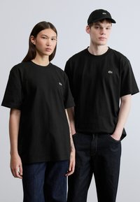 Δύο άτομα φορούν ίδια μαύρα βαμβακερά T-shirt με ένα μικρό πράσινο λογότυπο. Συνδυάζουν τα T-shirt με σκούρα τζιν παντελόνια.