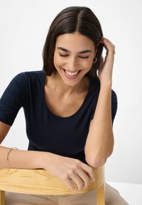 Marineblå kortærmet top med rund halsudskæring, glat tekstur; siddende stilling med hænderne hvilende på en træstol. Diskret detalje med et guldarmbånd.