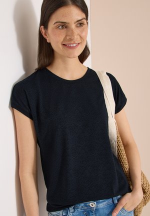 Young woman with dark hair wearing a black patterned t-shirt, blue jeans, and carrying a woven beige tote bag on her shoulder.