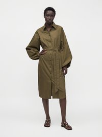 Personne portant une robe chemise ceinturée vert olive avec de longues manches bouffantes et des sandales noires, debout devant un fond blanc uni.