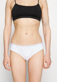 Fehér pamut bikini alsó, vékony gumis derékkal, amelyen ismétlődő Lacoste logó található. Simább tapintás és alacsony derekú kialakítás.