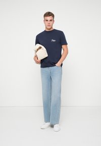 Joven con camiseta navy, jeans claros y zapatillas blancas sostiene una chaqueta beige con una mano en el bolsillo, de pie contra un fondo blanco.