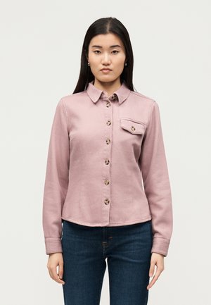 Woman standing wearing a dusty pink buttoned jacket with a single chest pocket and dark blue jeans against a plain light background.