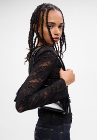 Zwarte lakleren handtas met een sleek ontwerp, voorzien van een ritssluiting en verstelbare schouderband, vastgehouden door een persoon in een kanten top.