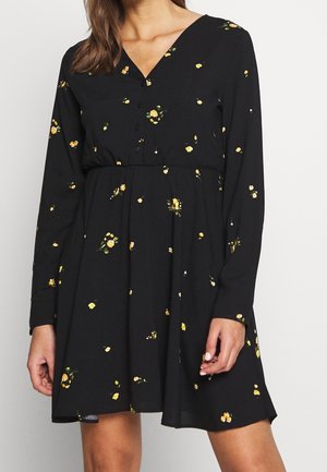 Robe noire à manches longues, décolleté en V, boutonnière à l'avant, et motif floral avec des fleurs jaunes sur un tissu texturé.