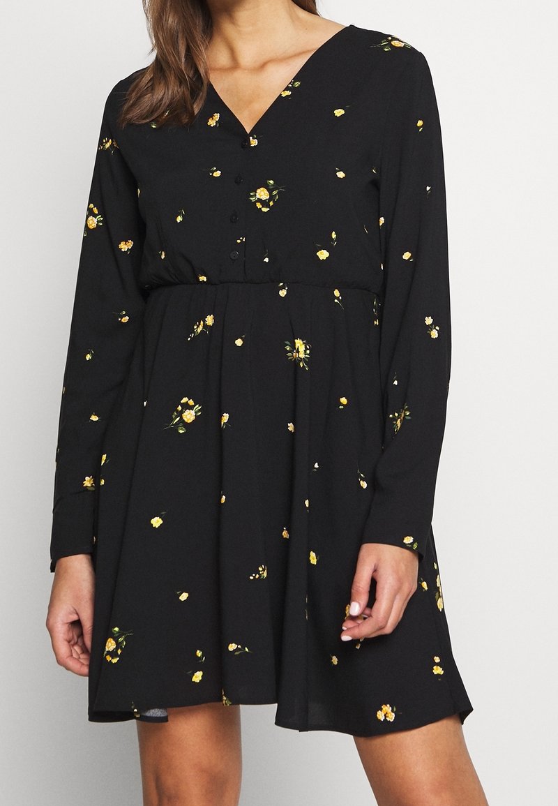 Schwarzes Kleid mit langen Ärmeln, V-Ausschnitt, Knopfleiste und floralen Muster, das gelbe Blumen auf einem strukturierten Stoff zeigt.