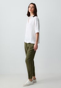 Weißes Leinenhemd mit kurzen Ärmeln und Knopfleiste; kombiniert mit olivgrünen Leinenhosen und weißen Sneakers. Schlichte, entspannte Silhouette.