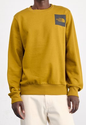 Mężczyzna noszący musztardowożółtą bluzę z okrągłym dekoltem i logo The North Face na lewej stronie klatki piersiowej, w zestawieniu z jasnobeżowymi spodniami.