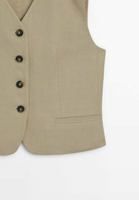 Veste beige sur mesure avec quatre boutons foncés et une poche passepoilée unique sur le côté droit sur fond blanc.