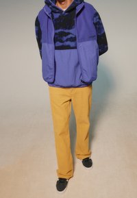 Chaleco de forro polar morado sobre una sudadera con capucha de patrón negro y azul, combinado con pantalones de pana amarillos y zapatillas negras. Diseño simple, corte informal.