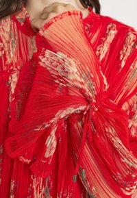 Punainen taitettu mekko, jossa on läpinäkyvä rakenne. Tässä on vaalean kerma- ja harmaita kukkakuvioita, nyöreillä varustetut kireät hihat ja röyhelökoristeet.