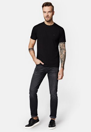 Czarny t-shirt z okrągłym dekoltem, dopasowany krój, w zestawieniu z ciemnymi dżinsami i czarnymi trampkami. Zauważalny tatuaż na przedramieniu, prosty i minimalistyczny wzór.