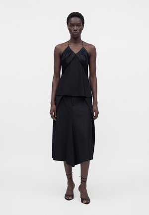 Femme portant un top dos nu noir, une jupe midi asymétrique noire et des sandales à talons à brides noires, debout devant un fond blanc.