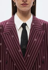 Oprijet blazer bordo barve z belo črtami, dvoredni kroj, kombiniran s črno kravato z značilnim kvadratnim logotipom in belo srajco.