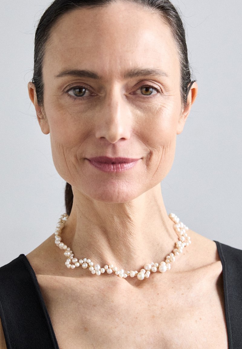 Κολιέ με μαργαριτάρια διαφόρων μεγεθών, διατεταγμένα περίτεχνα σε μια λεπτή αλυσίδα, φορεμένο πάνω από σκοτεινή μπλούζα.