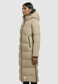 Longo cappotto puffer beige con cappuccio, caratterizzato da zip nere, un texture trapuntato, tasche laterali e un patch con logo nella parte inferiore.