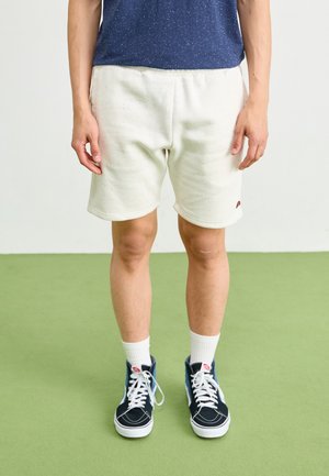 Shorts bianchi a macchie con una fascia elasticizzata in vita, abbinati a sneaker alte navy e calze bianche alla caviglia. In piedi su un pavimento verde.