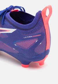 Fialové futbalové kopačky s textúrovaným sieťovaným zvrškom, kontrastnými ružovými akcentmi, posilneným pätovým okrajom a tvarovanou podrážkou s červenými kolíkmi.