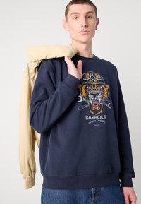 Navy sweatshirt featuring a tiger graphic with goggles and tools, ribbed cuffs, and a relaxed fit, paired with a beige jacket.
