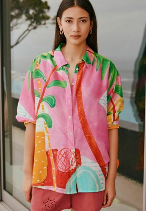 Young woman with straight dark hair wears oversized vibrant pink shirt with tropical leaf and abstract patterns, paired with red shorts.