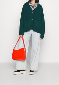 Zelený oversized cardigan s knoflíky, bílá a černá košile s puntíky pod ním, světlé denimové kalhoty s širokými nohavicemi a jasně červená kabelka přes rameno.
