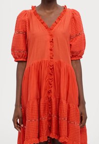 Czerwona bawełniana sukienka z bufiastymi rękawami, falbanami, frontem na guziki i warstwowym fasonem. Posiada teksturowane paski i koronkowe detale na rękawach.