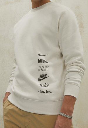 Osoba ubrana w biały sweter z kapturem z sześcioma różnymi czarnymi logo Nike ułożonymi pionowo z boku oraz beżowe spodnie.