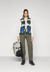 Barvna telovnik z modrim, belim in zelenim vzorcem, z dvema sprednjima žepoma, v kombinaciji z olivno zelenimi hlačami in črnimi pohodniškimi čevlji, s svetlim nahrbtnikom.