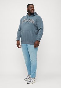 Grey hoodie with a front pocket and logo print, paired with light blue jeans and grey sneakers. Smooth fabric and relaxed fit.