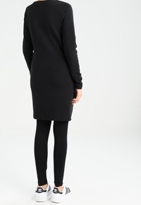 Robe noire à manches longues avec une coupe droite, accompagnée de leggings noirs slim et de baskets blanches avec des rayures noires.