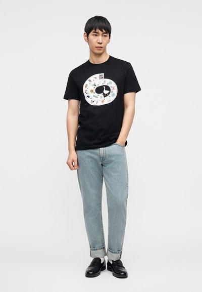 Μαύρο βαμβακερό μπλουζάκι με πολύχρωμο κυκλικό γραφικό σχέδιο με διάφορα εικονίδια. Το μοντέλο φοράει ανοιχτόχρωμο τζιν και μαύρα παπούτσια.