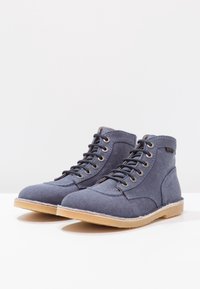 Bottines en toile bleue avec des lacets crème, bout rond et semelle en caoutchouc. Présente des détails de couture le long de la semelle et une étiquette de logo sur le côté.