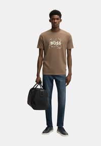 Camiseta marrón con un gráfico en blanco de "BOSS", combinada con pantalones vaqueros azules y zapatillas negras. Se sostiene un bolso de mano negro en una mano.